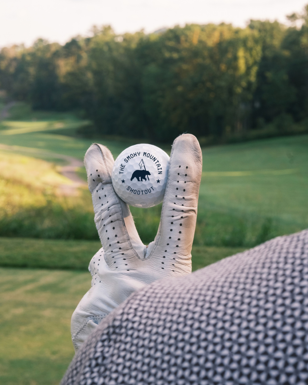 Gloved hand holding a Smoky Mountain Shootout golf ball on a green fairway